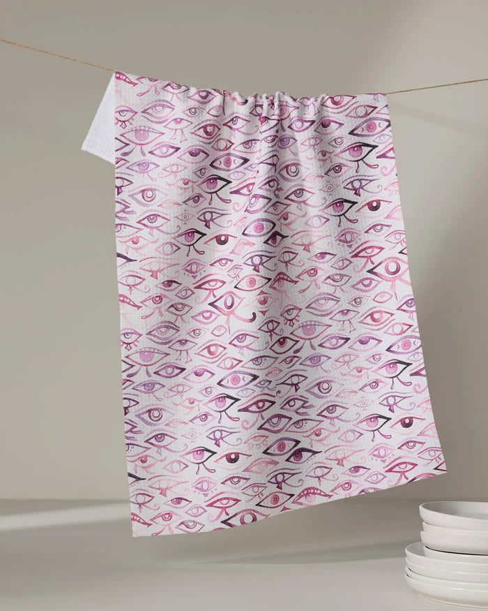 The Eye Of Horus tea towel by Maayan Alper-Swan, featuring a quick-drying fabric and purple-pink illustrated eye pattern, hangs on a string above a stack of white plates.