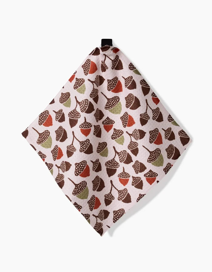 The Kayla Ann Fall Abundance dishcloth, crafted from Geoweave fabric, showcases brown, red, and green acorns and leaves on a light background and hangs from a black loop against a white surface.