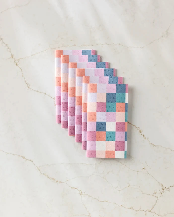 Seven pastel-colored "Fall Checkers" envelopes by Claire Iglesias, featuring a pixelated square pattern, are fanned out on a light marble surface, offering soft hues and sustainable appeal.