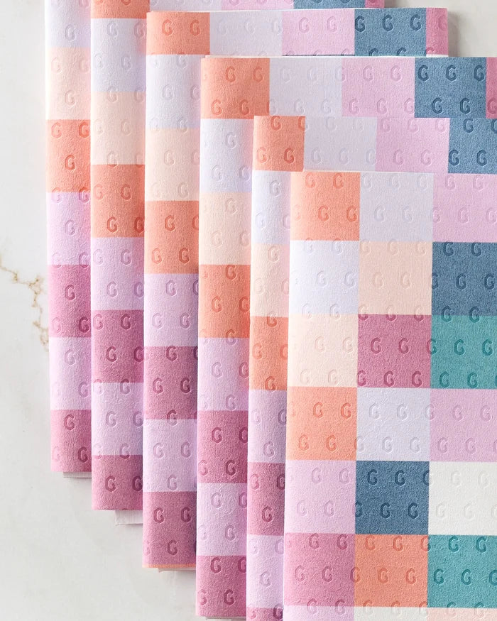 Five folded, reusable "Fall Checkers" paper towels by Claire Iglesias, featuring a pastel checkerboard in pink, peach, lavender, and teal with embossed Gs—arranged diagonally on marble—offer a stylish swap for disposables.