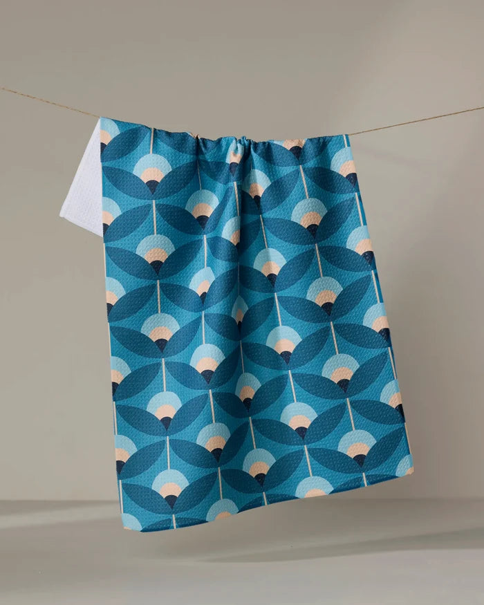 The Blissful Flamingo "Fall Florals" tea towel, featuring a quick-drying fabric and blue floral pattern with beige accents, hangs on a clothesline against a plain, neutral background.