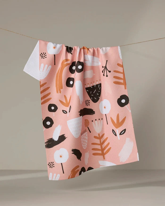 The "Fall Flowerbed" tea towel by Marina Solodka features a peach hue with playful abstract white, black, and brown floral and leaf patterns, shown hanging on a clothesline against a neutral backdrop.