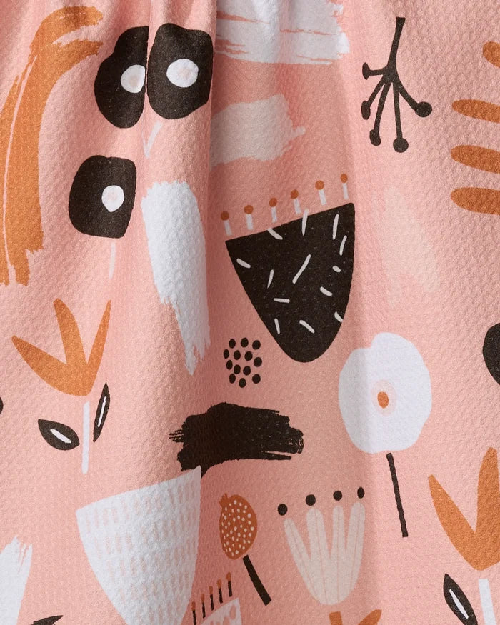 The Fall Flowerbed recycled kitchen towel by Marina Solodka features a pink backdrop with black, white, and orange shapes—flowers, dots, leaves, and brushstrokes—for a playful, modern touch.