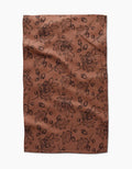 The Fall Foliage kitchen towel by Aubrey Fairchild features eco-friendly brown fabric with a black oak leaf and acorn pattern, creating an autumnal, botanical vibe. Laid flat with slight wrinkles, it makes a charming addition to any kitchen.