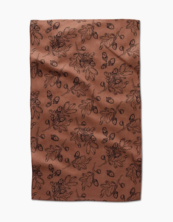 The Fall Foliage kitchen towel by Aubrey Fairchild features eco-friendly brown fabric with a black oak leaf and acorn pattern, creating an autumnal, botanical vibe. Laid flat with slight wrinkles, it makes a charming addition to any kitchen.