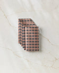 Six folded Fall Gingham cloth napkins by GEOMETRY, featuring a brown and black checkered pattern, are neatly stacked on a white marble surface—an eco-friendly alternative to traditional paper towels.