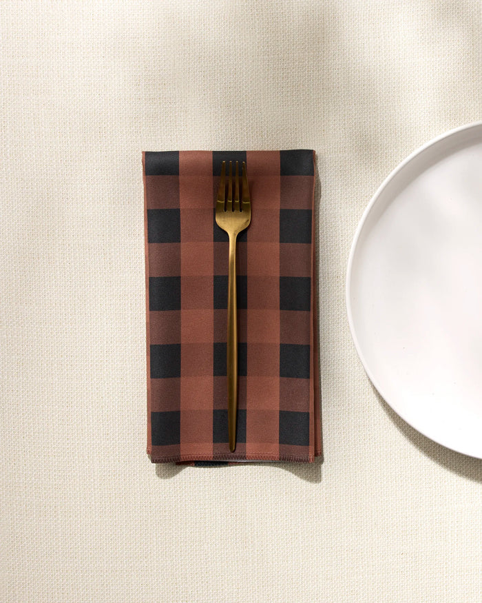 A gold fork rests on a folded GEOMETRY Fall Gingham reusable napkin, placed on a textured cream-colored tablecloth beside a white plate near the right edge.
