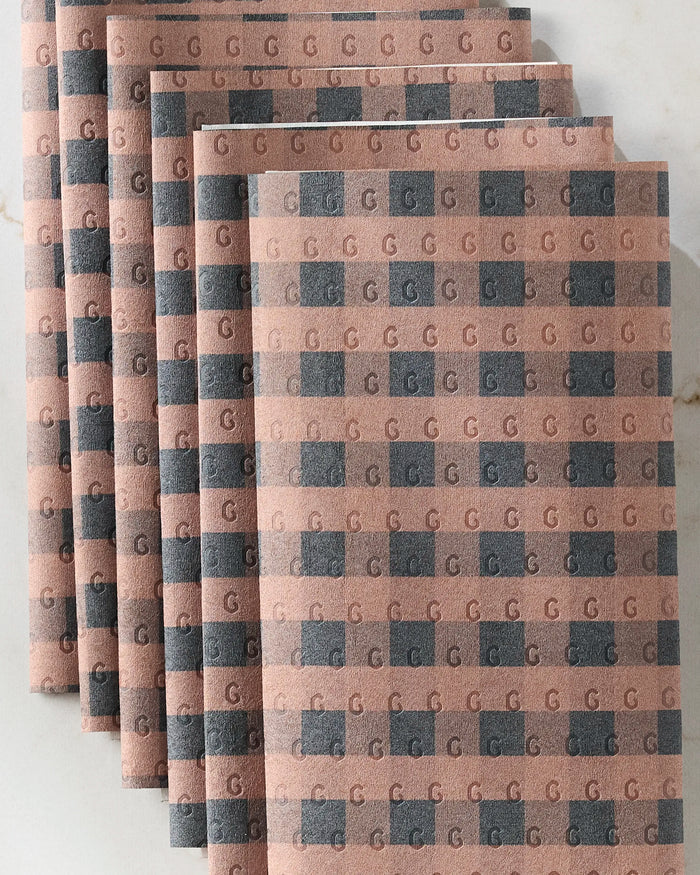 Six Fall Gingham napkins by GEOMETRY, featuring a geometric pattern of alternating gray and tan squares with repeating "G" designs—an elegant, eco-friendly kitchen towel alternative displayed on a light surface.