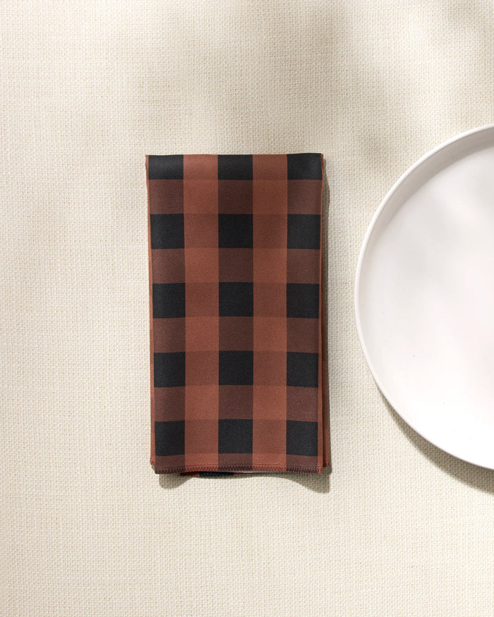 A GEOMETRY Fall Gingham folded reusable napkin with a brown and black checkered pattern rests on a light textured surface beside the edge of a plain white plate.
