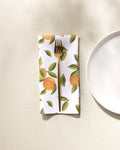 A gold fork rests on a GEOMETRY Fallbrook Oranges absorbent cloth napkin featuring an orange and green leaf pattern, placed beside a white plate on a light textured surface.