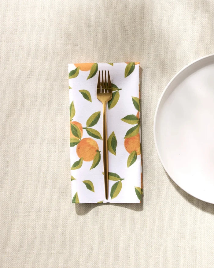 A gold fork rests on a GEOMETRY Fallbrook Oranges absorbent cloth napkin featuring an orange and green leaf pattern, placed beside a white plate on a light textured surface.