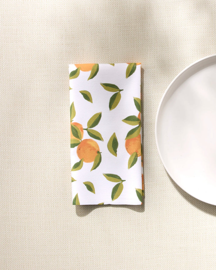 A folded white GEOMETRY Fallbrook Oranges napkin, featuring an orange fruit and green leaf pattern, rests on a light textured surface by a plain white plate—part of an absorbent dinner napkin set.
