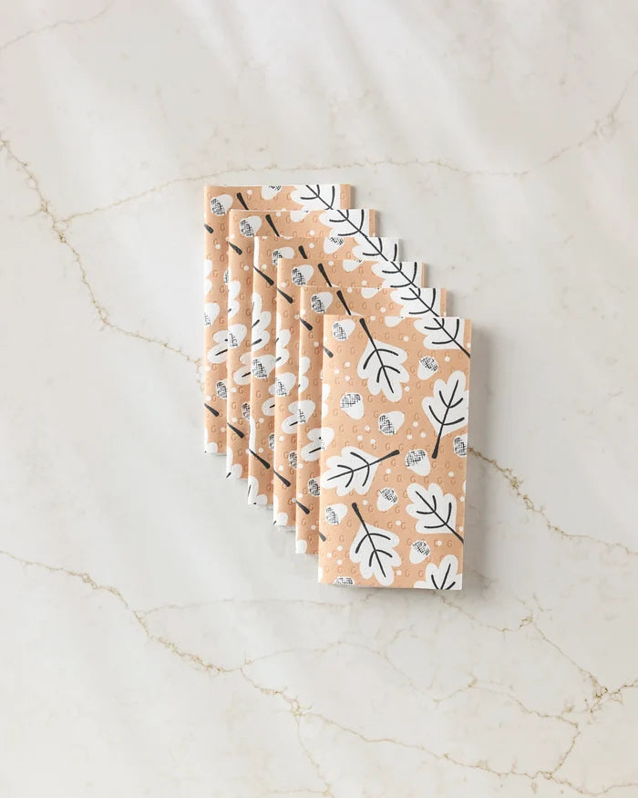 Six Falling Acorns rectangular bars by Charlie Rowan Designs are wrapped in peach, eco-friendly paper towels featuring a white and black leaf pattern, neatly displayed on a light, marble-patterned surface.
