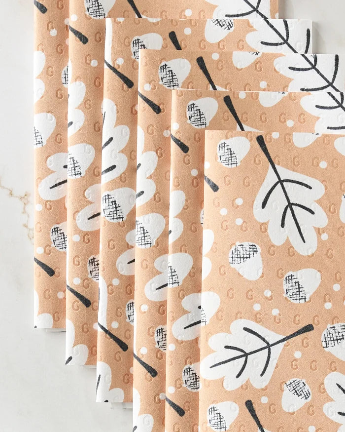 Five folded reusable paper towels from Charlie Rowan Designs, in the Falling Acorns print with tan background and white and black leaves and dots, are arranged fan-shaped on a light marble surface.