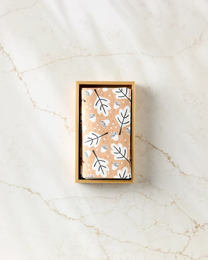 A rectangular box with a lid from Charlie Rowan Designs, containing Falling Acorns, wrapped in peach eco-friendly towels with white leaves and animal faces, rests on a white marble surface.