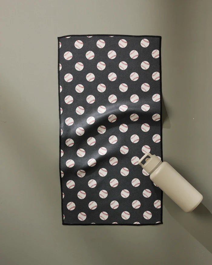 The Geometry Fastball towel, featuring a black design with a repeating baseball pattern, lies flat on a light gray surface with a beige water bottle at its lower right corner. Ultra-absorbent and fast drying, it's ideal for workouts or daily use.