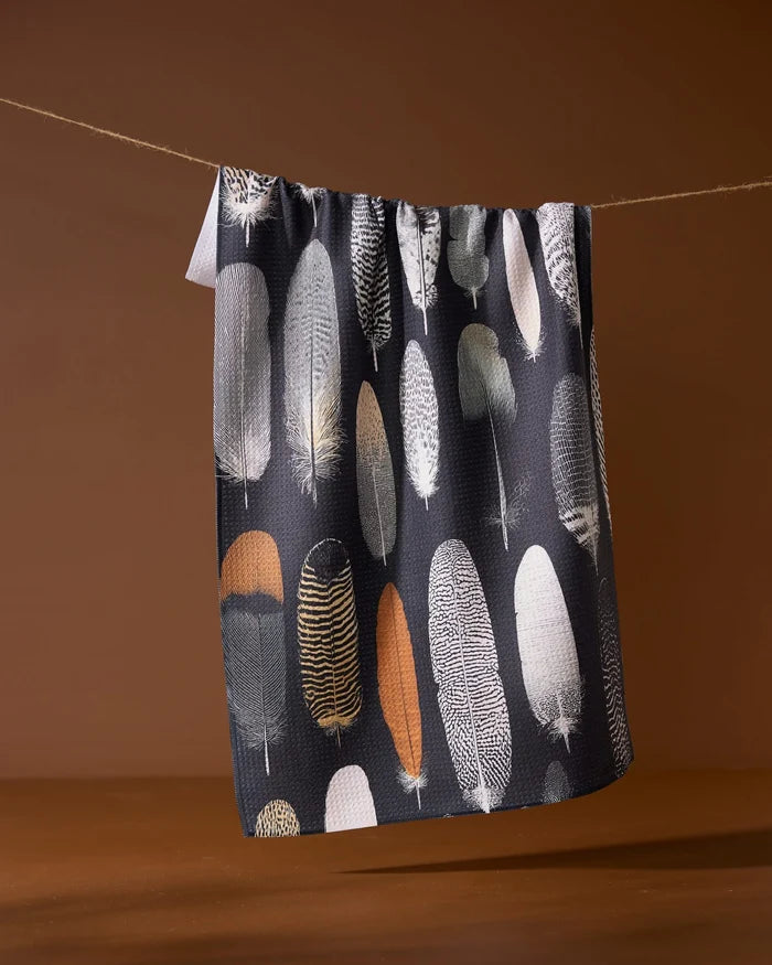 The Geometry Featherfall towel, an eco-friendly, dark-toned piece featuring neutral feather patterns, hangs on a line set against a brown background.