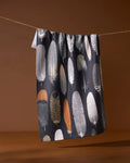 The Geometry Featherfall towel, an eco-friendly, dark-toned piece featuring neutral feather patterns, hangs on a line set against a brown background.