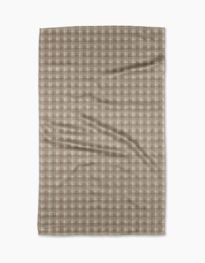 A Geometry Feelin Sandy tea towel, featuring a beige and white checkered pattern in Geoweave fabric, lies flat on a white background with gentle creases visible across its surface.