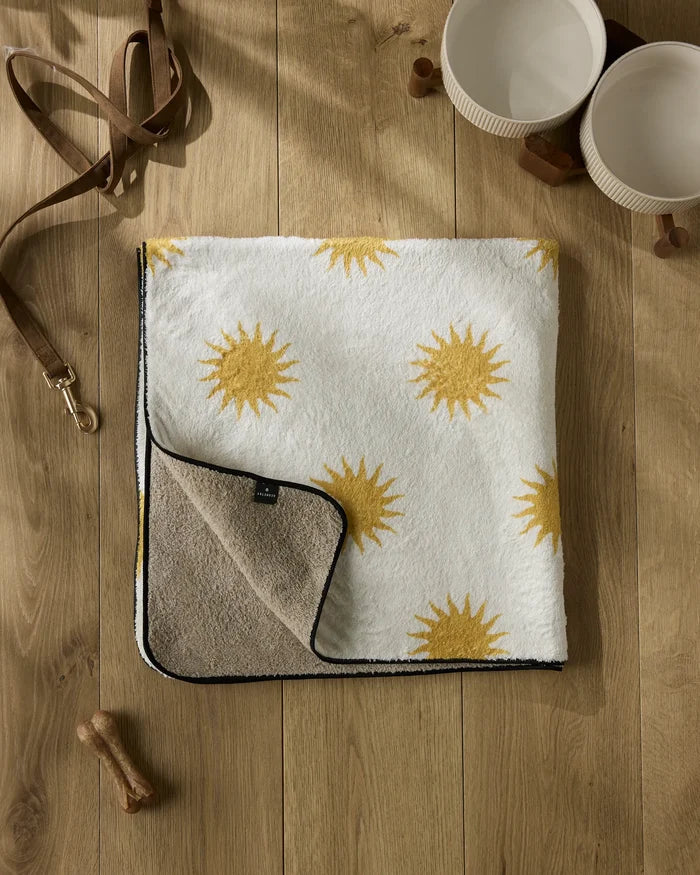 The Geometry Feeling Sunny plush dog towel with yellow sun patterns lies folded on a wooden floor, alongside two cream pet bowls, a brown dog leash, and some treats. The towel’s underside is beige.