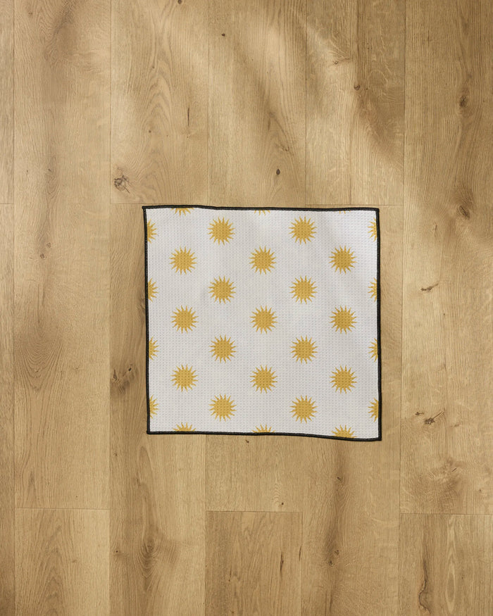 The Geometry Feeling Sunny paw towel, featuring a square shape with a repeating yellow sun pattern, black border, and white background, is laid flat on a light-colored wooden floor.