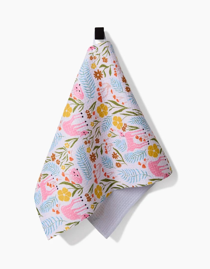 The Fern and Fawn bar towel by Kayla Ann features pink animals, orange and yellow flowers, plus green and blue leaves on white Geoweave fabric. It hangs from a black loop against a white background.