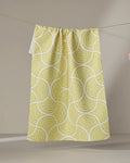 The Dabney Lee "Ferns" tea towel, made from recycled materials, displays a yellow and white repeating fan pattern. Quick-drying and stylish, it hangs from a clothesline against a neutral background for a sustainable touch.