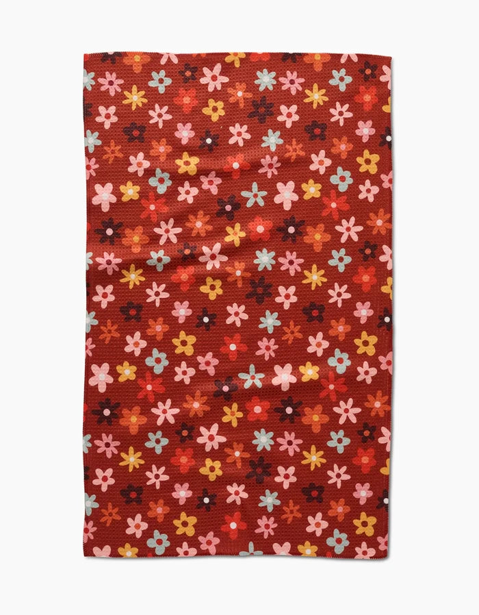 The Sara Mari Field Of Fall Daisies is a rectangular red tea towel with a pattern of small, colorful flowers in pink, yellow, blue, and orange evenly distributed across the surface.