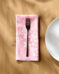 A fork rests on a Hufton Studio "Field of Flowers" pink floral-patterned reusable cloth napkin, neatly folded on a light wooden table beside a white plate as sunlight casts soft shadows nearby.