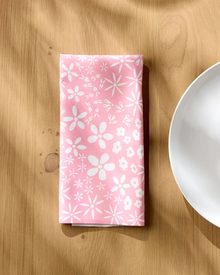 A Field of Flowers pink dinner napkin from Hufton Studio, with white floral patterns, is neatly folded on a light wooden table beside a white ceramic plate.