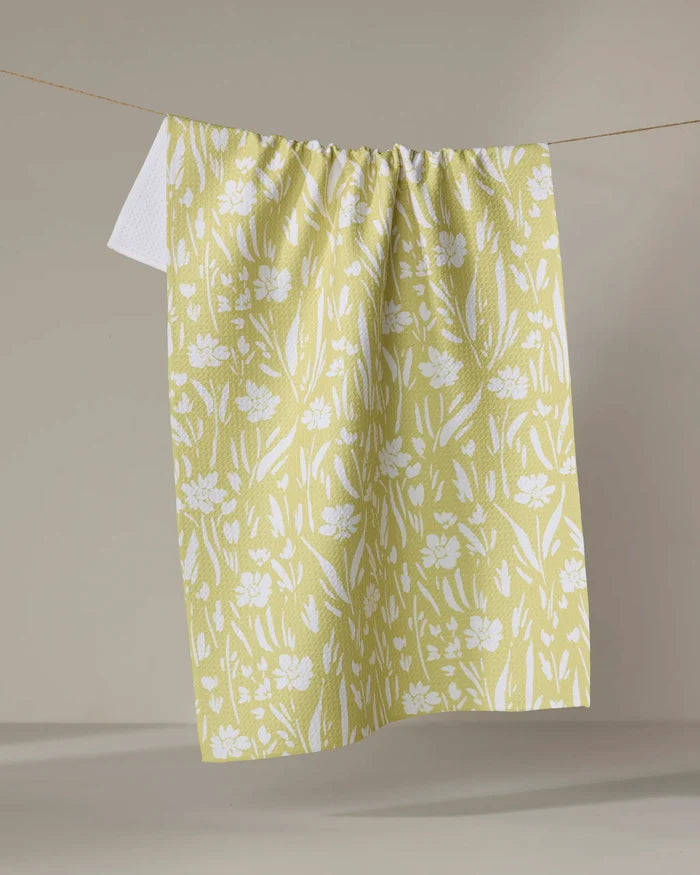 The "Field of Wildflowers" tea towel by Ashley Celena, featuring a white floral pattern on yellow and made from recycled materials, hangs on a clothesline against a neutral background.