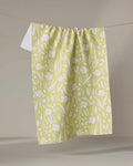 The "Field of Wildflowers" tea towel by Ashley Celena, featuring a white floral pattern on yellow and made from recycled materials, hangs on a clothesline against a neutral background.