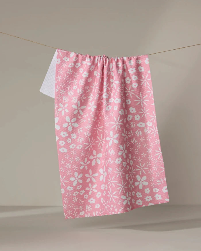 The Fields of Flowers tea towel by Hufton Studio, featuring a pink hue and white floral pattern made from recycled materials, hangs on a clothesline against a beige background with another white cloth partially visible behind it.