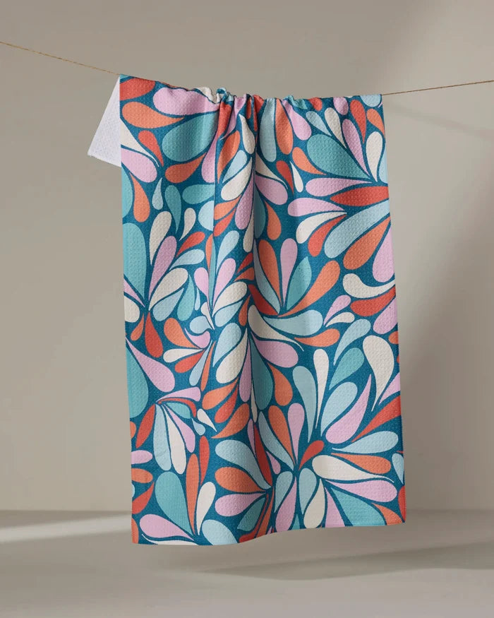 The "First Flight" kitchen towel by Rebecca Woolbright features a bold, abstract floral pattern in blue, pink, orange, and white and hangs on a clothesline against a neutral background.