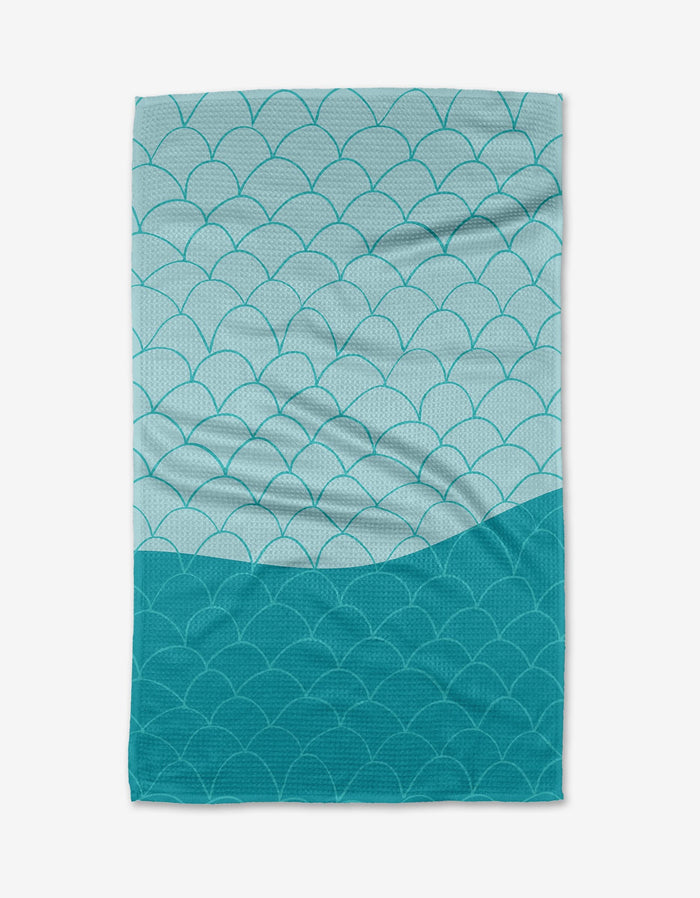 The Fish Scales tea towel by Marta Harvey is a rectangular towel made from recycled materials, boasting an overlapping blue and teal scallop design—darker teal at the bottom, lighter blue at the top.