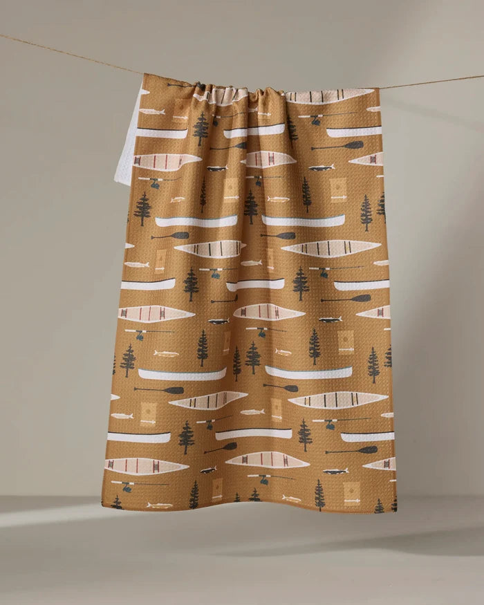The Coit Creative “Fishing On the Lake” tea towel features an ultra-absorbent brown background with white, black, and beige canoes, paddles, trees, and geometric shapes. It hangs neatly on a line.