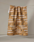 The Coit Creative “Fishing On the Lake” tea towel features an ultra-absorbent brown background with white, black, and beige canoes, paddles, trees, and geometric shapes. It hangs neatly on a line.