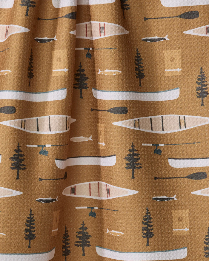 The "Fishing On the Lake" recycled kitchen towel by Coit Creative features a brown background with a repeating pattern of canoes, paddles, pine trees, fish, and camp lanterns in white, black, blue, and green.