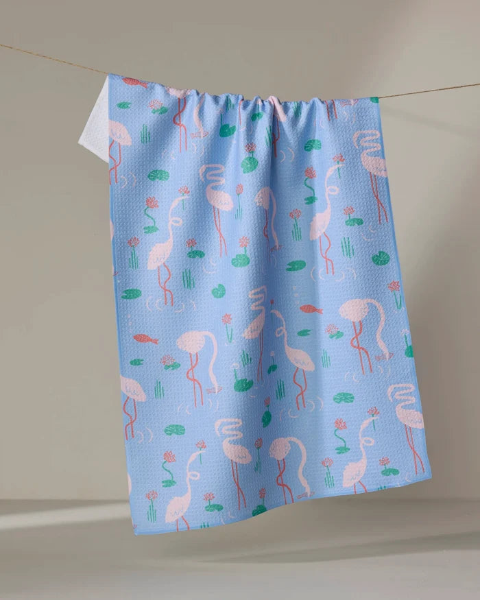 The Flirty Flamingo tea towel by Elisabeth Wing is light blue, made from recycled materials, and features pink flamingos, green lily pads, and small pink flowers hanging against a neutral background.