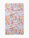 The Floppsie tea towel by Amy MacCready features a quick-drying fabric with a whimsical, repetitive floral pattern of red, yellow, and purple flowers with green stems and leaves on a light pink background.