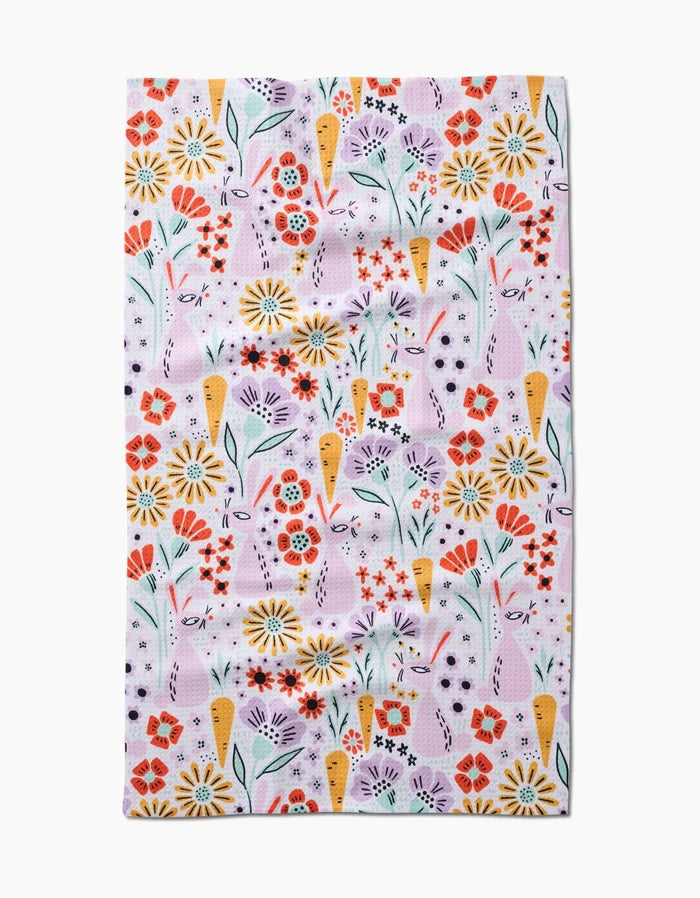 The Floppsie tea towel by Amy MacCready features a quick-drying fabric with a whimsical, repetitive floral pattern of red, yellow, and purple flowers with green stems and leaves on a light pink background.