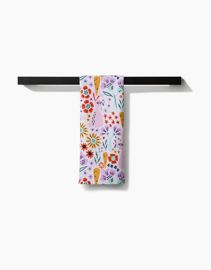 The Floppsie tea towel by Amy MacCready features a floral and carrot pattern with red, yellow, and orange flowers. Made from quick-drying recycled materials, it hangs neatly over a black towel bar against a white background.