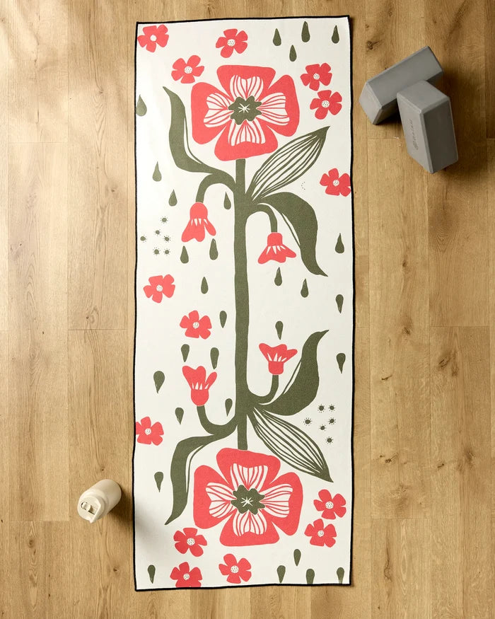 The Ash Cascade Flora Stamp yoga mat with a bold pink and green floral design is spread on a wooden floor, accompanied by a white water bottle below and a silicone grip towel plus two gray yoga blocks at the top right.