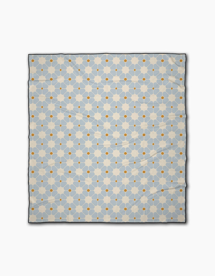 The Geometry Flora Tile Light Blue is a quick-drying beach blanket with a repeating geometric pattern of light blue hexagons and small orange circles on a white background.