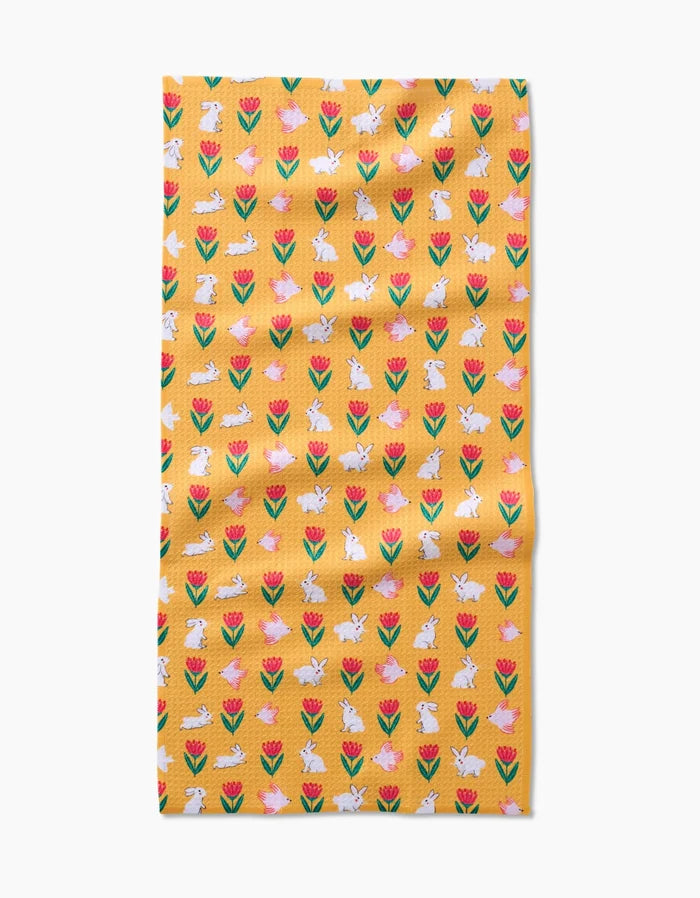 The Floral Bunny Parade kitchen towel by Ceyda Alasar features a yellow recycled fabric with red and pink tulips and white bunnies in a repeating pattern, shown slightly wrinkled on a white background.