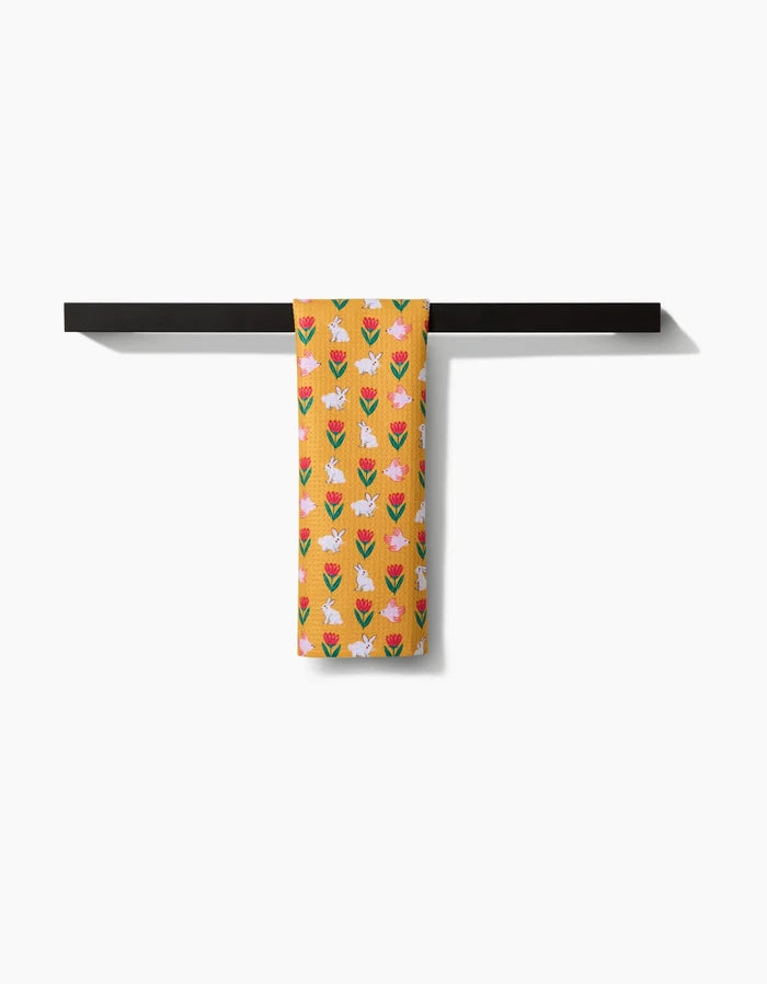 The Floral Bunny Parade towel by Ceyda Alasar, a yellow ultra-absorbent piece with pink, white, and blue tulip patterns, hangs neatly over a black horizontal bar against a plain white background.