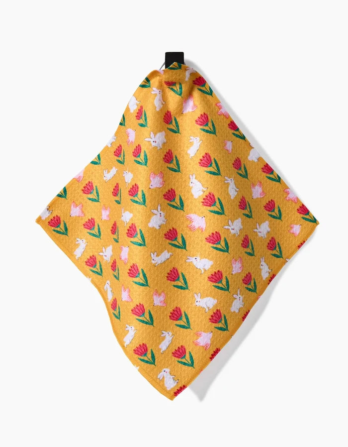 Floral Easter Parade by Ceyda Alasar is a yellow cloth made from recycled materials, featuring white bunnies and pink tulips with green leaves. It hangs from a black loop against a white background.