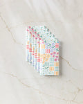 Six "Floral Garden" cloth napkins by Charlie Rowan Designs, featuring vibrant floral and geometric patterns inspired by reusable paper towels, are neatly fanned out on a white marble surface.
