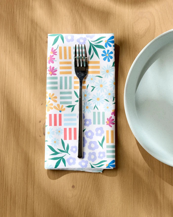 A silver fork rests on Charlie Rowan Designs’ Floral Garden eco-friendly napkin beside a white ceramic plate on a sunlit wooden table.