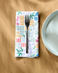A silver fork rests on Charlie Rowan Designs’ Floral Garden eco-friendly napkin beside a white ceramic plate on a sunlit wooden table.